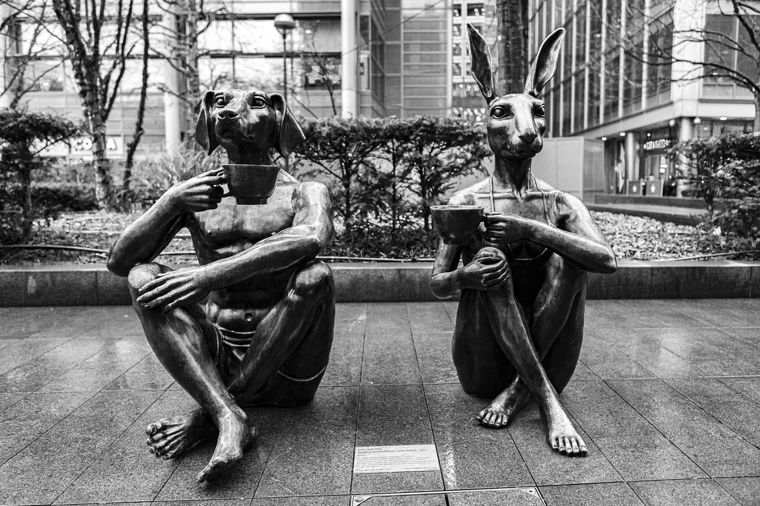 Two statues of people sitting side by side drinking coffee, but the male statue on the left has a dog's head and the female statue on the left has a rabbit head.