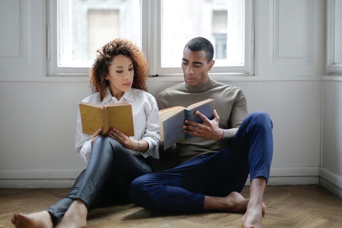 Woman and man sitting on the floor each reading a book. The man is pointing to a part of his book he wants the woman to see.