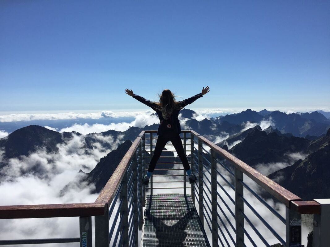 Person standing on a fenced bridge that looks over the tops of mountains.