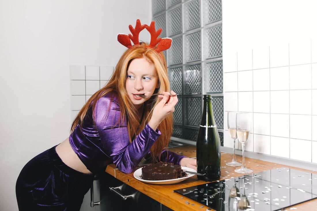 Attractive woman wearing holiday reindeer horns leaning over a counter and eating small forkfuls of chocolate cake.